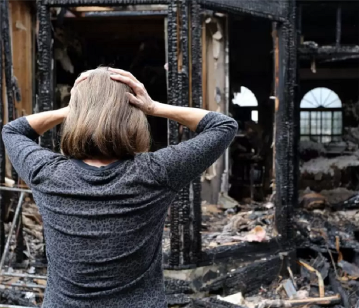 Woman facing fire damaged home with palms pressed to sides of head in state of worry